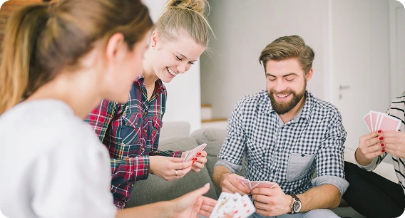 Drie vrienden zitten op een bank te kaarten, glimlachend en betrokken. De sfeer is ontspannen en vrolijk, met casual kleding en een lichte omgeving.