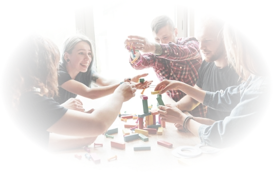 Een groep van vijf mensen, glimlachend en betrokken, werkt samen om een kleurrijke blokkentoren te bouwen aan een tafel. Het zonlicht stroomt door een raam naar binnen en creëert een warme sfeer.