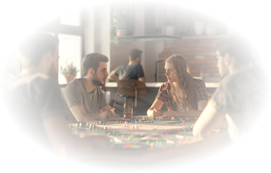 Een groep jonge volwassenen zit rond een tafel, intens geconcentreerd op een bordspel. Zonlicht valt door de ramen naar binnen in de gezellige kamer met bakstenen muren, waardoor een warme sfeer ontstaat.