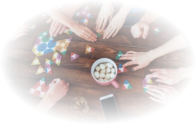 Verschillende handen spelen met kleurrijke driehoekige puzzelstukjes op een houten tafel. In de buurt liggen een smartphone, snacks, een gamecontroller en drankjes, wat een levendige, sociale sfeer creëert.
