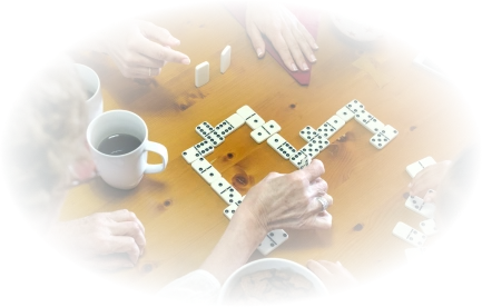 Een groep mensen speelt domino op een houten tafel. Kopjes koffie, schalen met snacks, een smartphone en een fidget spinner liggen verspreid, wat een gezellige, sociale sfeer creëert.