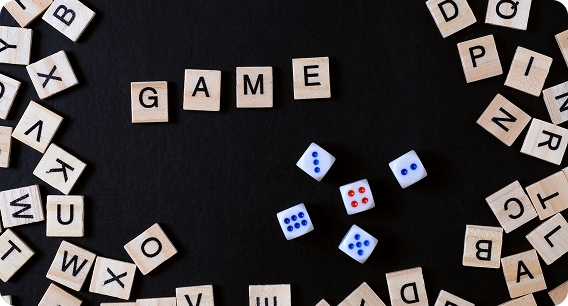 Wooden letter tiles spell "GAME" in the center on a black background, surrounded by scattered tiles and four dice showing various numbers.